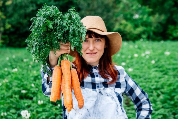 Portrait of a beautiful woman farmer holds a bunch of carrots in a straw hat and surrounded by the many plants in her vegetable garden. - Foto, afbeelding