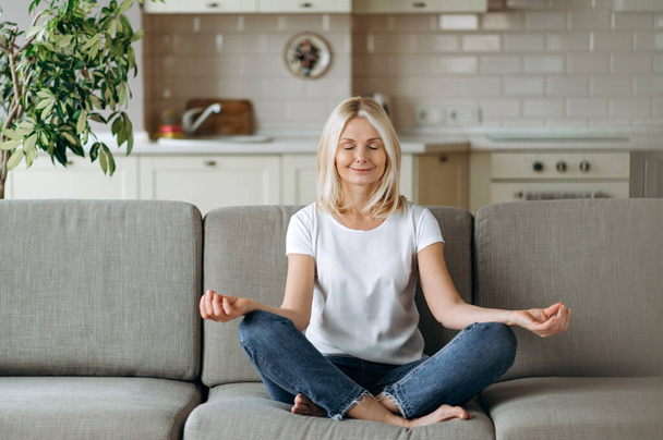 Satisfied attractive middle aged female sits on sofa at home and chilling. Blonde mature is practise yoga and meditation in lotus position with eyes closed, relieve stress, calmness concept - Фото, зображення
