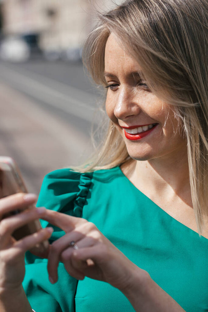 beautiful young smiling blonde woman with red lipstick and in a green dress photographs herself on a mobile phone - Fotoğraf, Görsel