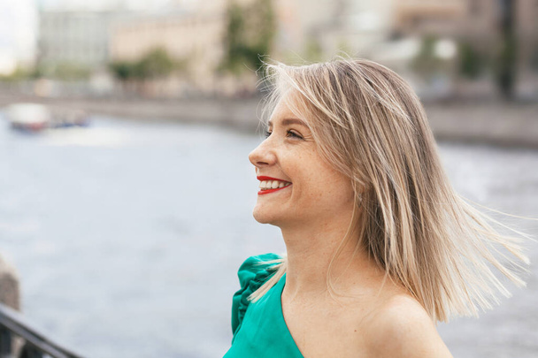 portrait of a beautiful smiling young woman with red lipstick and in a green dress on the background of the river embankment in the city - Zdjęcie, obraz