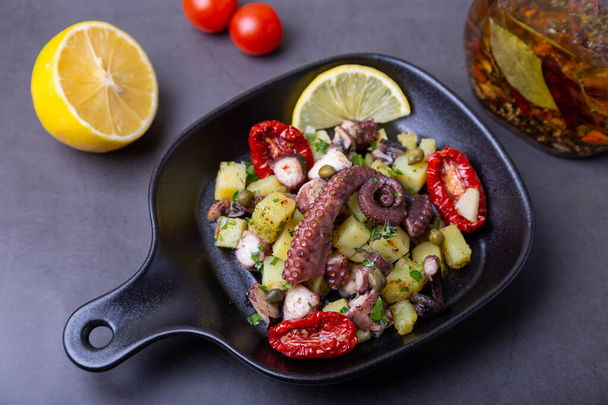 Warm salad with octopus, potatoes, sun-dried tomatoes, capers and lemon in a black small pan. Close-up, black background. - Photo, image