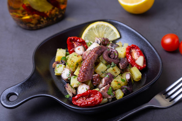 Warm salad with octopus, potatoes, sun-dried tomatoes, garlic and lemon in a black small pan. Close-up, black background. - Fotografie, Obrázek