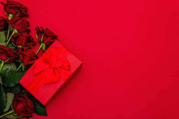マットな赤のファミランの背景に新鮮なバーガンディのバラとギフトボックスの花束。香り高い赤い花、バレンタインデーのギフトコンセプト、結婚式や誕生日、トップビュー - 写真・画像