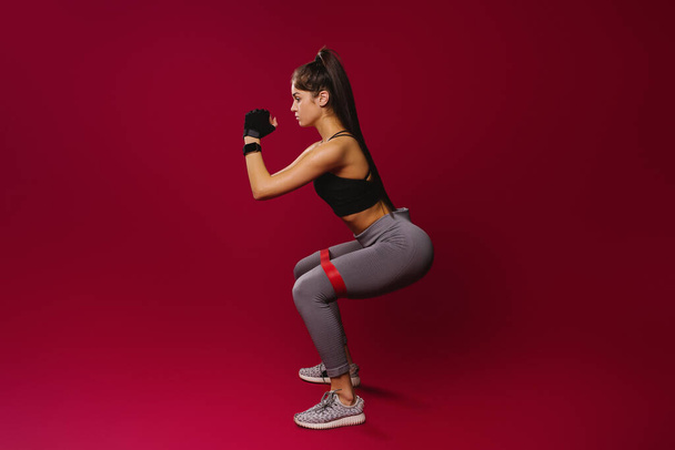 Slim girl in sportswear with elastic bandage doing exercises on a red background with empty side space. The concept of a healthy lifestyle and a beautiful figure. - Foto, imagen