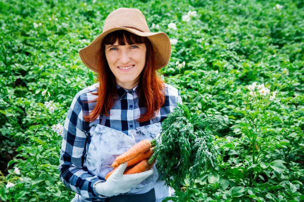 Porträt einer schönen Bäuerin, die einen Strauß Möhren im Strohhut hält und von den vielen Pflanzen in ihrem Gemüsegarten umgeben ist. - Foto, Bild
