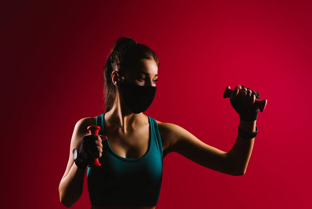 Retrato de uma menina de esportes olhando para o lado em uma pandemia têxtil máscara preta e halteres em suas mãos. - Foto, Imagem