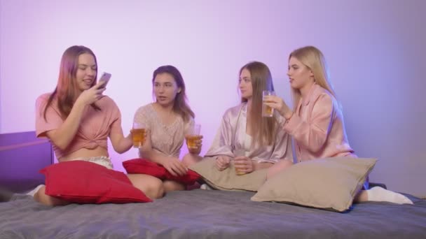 Четыре счастливые молодые женщины в пижаме сидят на кровати. Одна женщина записывает видео на телефон с тостами и стаканами на девичнике - Кадры, видео