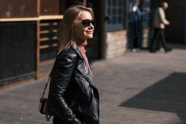 young beautiful smiling stylish blonde woman in sunglasses, black pants and leather jacket posing on the don of buildings in an urban setting - Photo, image