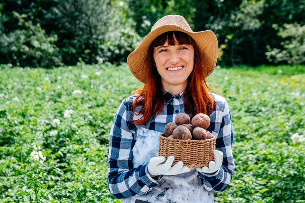 Portrait beautiful female farmer holds a potatoes in a wicker basket wearing a straw hat and surrounded by the many plants in her vegetable garden. - Photo, Image