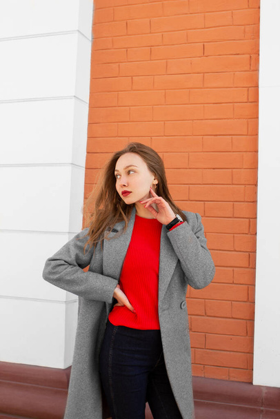 コートの茶色の髪の若い女性は、手に時計と赤いシャツ、通りに立って、赤レンガの壁の前で左側を見て。白人女性。都会的なスタイルとファッション。普段着 - 写真・画像