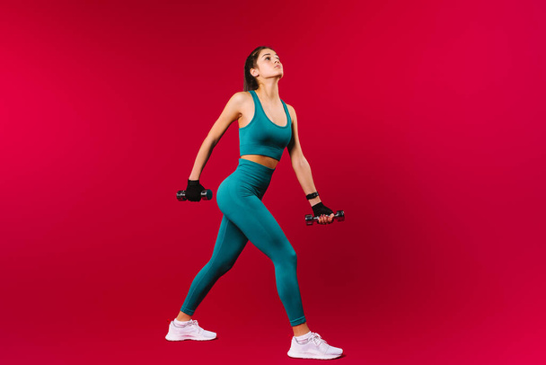 A girl with a slender and pumped-up body. In sportswear with dumbbells in his hands looks up on a red background with side space. - Photo, Image