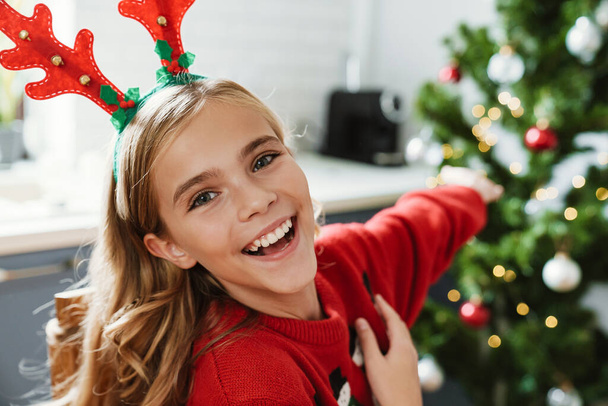 Happy caucasian girl wearing toy deer horns smiling and showing Christmas tree at home - Foto, imagen