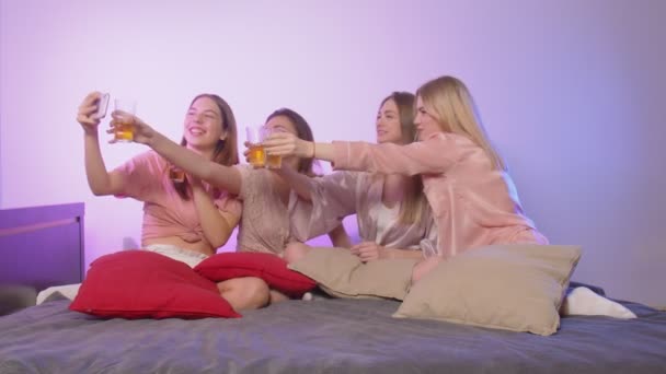 Vier glückliche hübsche junge Frauen im Schlafanzug und mit Brille sitzen auf dem Bett und nehmen Video-Glückwünsche auf - Filmmaterial, Video