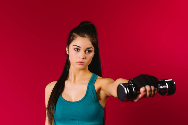 A serious sports girl holds a dumbbell on her outstretched hand in front of her on a red background with side space. Sport and healthy lifestyle concept. - Фото, зображення