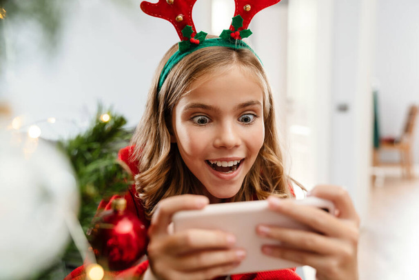 Excited caucasian girl wearing toy deer horns playing video game on cellphone at home - Foto, Bild