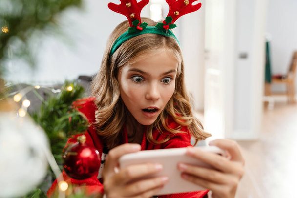Shocked caucasian girl wearing toy deer horns playing video game on cellphone at home - Foto, Imagem
