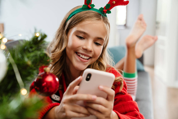 Happy caucasian girl wearing toy deer horns smiling and using cellphone at home - Zdjęcie, obraz