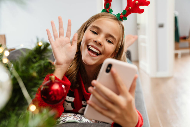 Happy caucasian girl wearing toy deer horns waving hand and using cellphone at home - Fotoğraf, Görsel