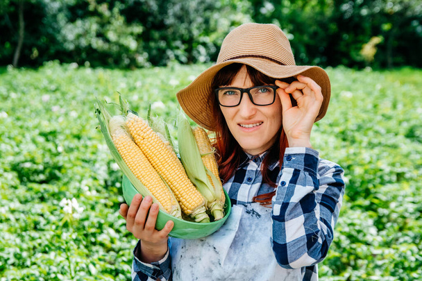 Retrato de una hermosa agricultora sosteniendo una mazorca de maíz en un plato, sonriendo a la cámara con un sombrero de paja y rodeada de muchas plantas en su huerto. - Foto, Imagen