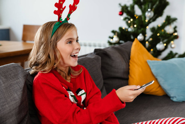 Ευτυχισμένο ωραίο κορίτσι φορώντας κέρατα ελαφιού παιχνίδι βλέποντας τηλεόραση και χρησιμοποιώντας τηλεχειριστήριο στο σπίτι - Φωτογραφία, εικόνα