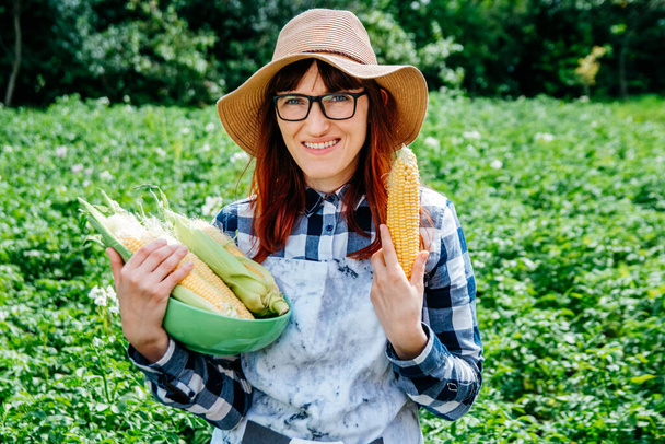 Porträt einer schönen Bäuerin, die einen Maiskolben in einem Teller hält, mit einem Strohhut in die Kamera lächelt und von vielen Pflanzen in ihrem Gemüsegarten umgeben ist. - Foto, Bild