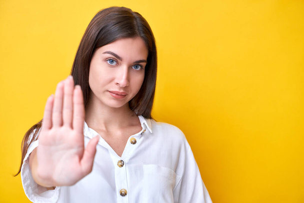 Brunette girl showing stop gesture with hands, looking at the camera with strong face isolated on yellow background, keep distance - Foto, imagen