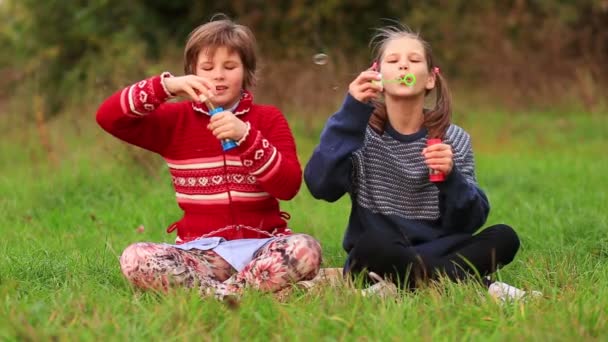 Cute child girls sits on grass with crossed legs and blowing soap bubbles in park. Playing with soap bubbles - Footage, Video