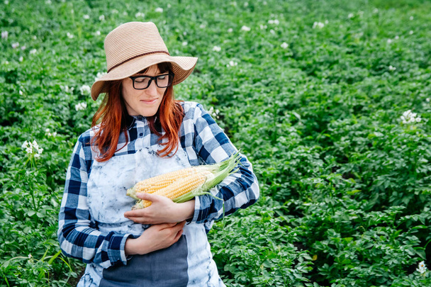 Portret pięknej farmerki trzymającej kolby kukurydzy w słomkowym kapeluszu i otoczonej mnóstwem roślin w ogrodzie warzywnym. - Zdjęcie, obraz
