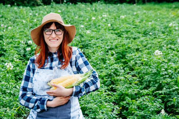 Portret pięknej farmerki trzymającej kolby kukurydzy, uśmiechającej się do kamery w słomianym kapeluszu i otoczonej mnóstwem roślin w ogrodzie warzywnym. - Zdjęcie, obraz