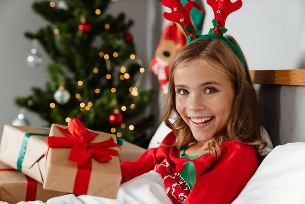 Veselý šťastný dívka na sobě hračky jeleni rohy držení vánoční dárek při ležení v posteli - Fotografie, Obrázek