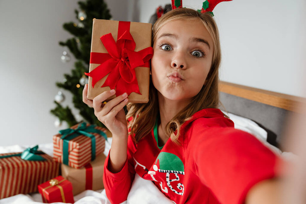 Cheerful caucasian girl wearing toy deer horns taking selfie on cellphone while lying in bed, holding gift box - Φωτογραφία, εικόνα