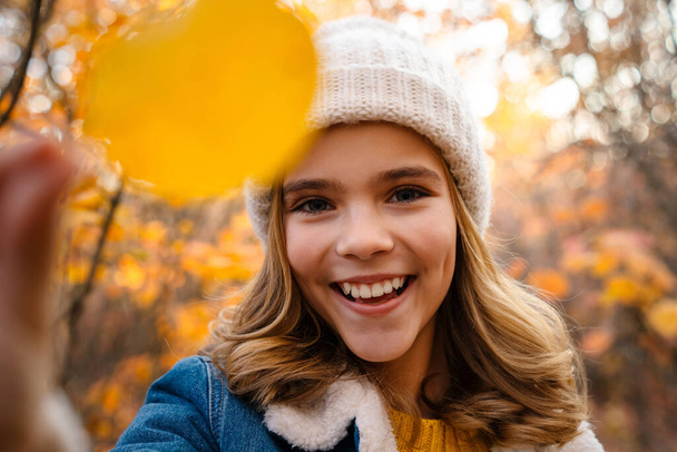 Image of young pretty optimistic girl walking outdoors in autumn park with leaf in hand - Photo, Image