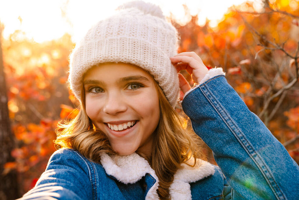 Image of young pretty happy girl walking outdoors in autumn park taking a selfie by camera - Fotó, kép