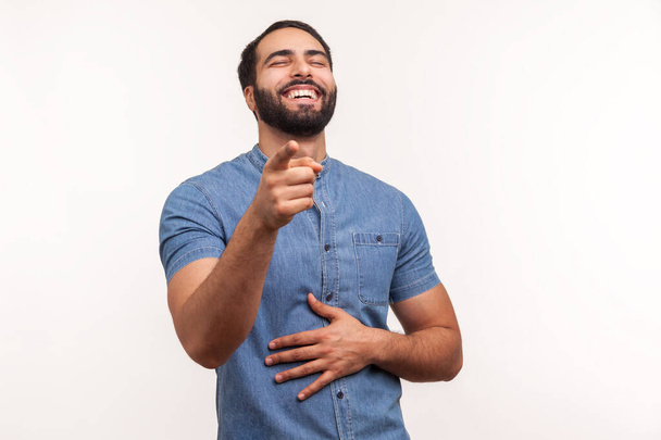 Extremely happy bearded man laughing holding hand on belly and pointing finger at you looking at camera, funny ridiculous joke. Indoor studio shot isolated on white background - Foto, afbeelding