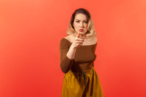 Oh my God, I can't believe it's you. Portrait of confused serious angry woman with blonde hair in casual clothes pointing at the camera with finger. Indoor studio shot isolated on red background - Foto, imagen