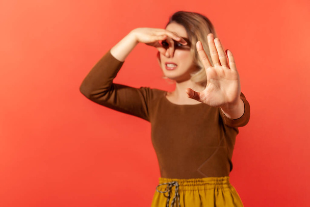 Portrait of displeased woman grimacing with disgust, holding breath, pinching nose with fingers to avoid bad smell and showing stop gesture. Indoor studio shot isolated on red background - Fotó, kép