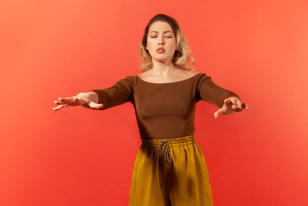I don't see anything. Portrait of blonde woman standing with closed eyes and outstretched hands, trying to find obstacles while walking blind. Indoor studio shot isolated on red background - Photo, image