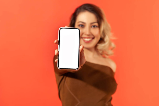 Positive smiling woman holding and showing smartphone with empty white display, freespace for your advertisement, tariffs and bonuses, mobile app. Indoor studio shot isolated on orange background - Fotoğraf, Görsel