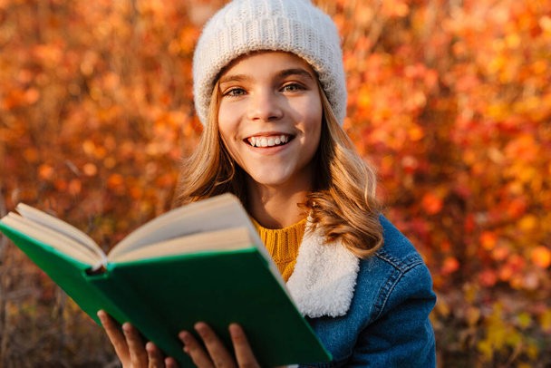 Image of young optimistic girl sitting outdoors in autumn park and reading book - Valokuva, kuva