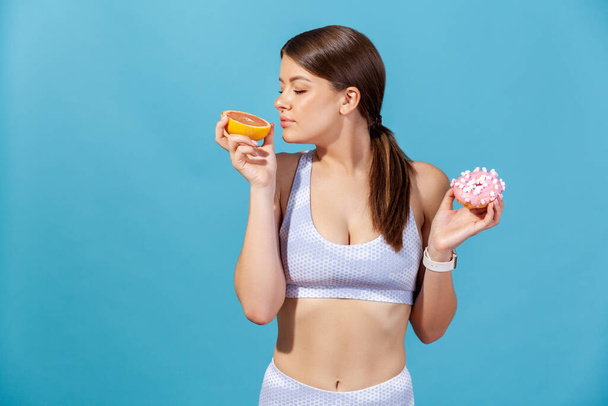 Sportive woman in white top holding in hands half of juicy grapefruit and round donut with pink icing, enjoying fresh aroma of ripe fruit. Indoor studio shot isolated on blue background - Foto, immagini