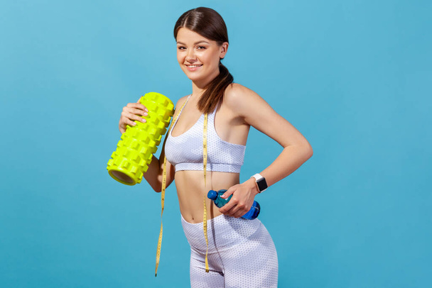 Profile portrait smiling athletic woman in white sportive top and tights holding bottle with spring water and roller massager, doing fitness exercises. Indoor studio shot isolated on blue background - Fotoğraf, Görsel
