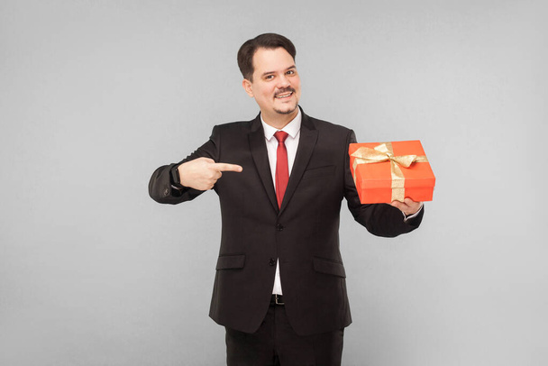 Business man holding red gift box. Pointing finger. Indoor, studio shot, isolated on gray background - Valokuva, kuva