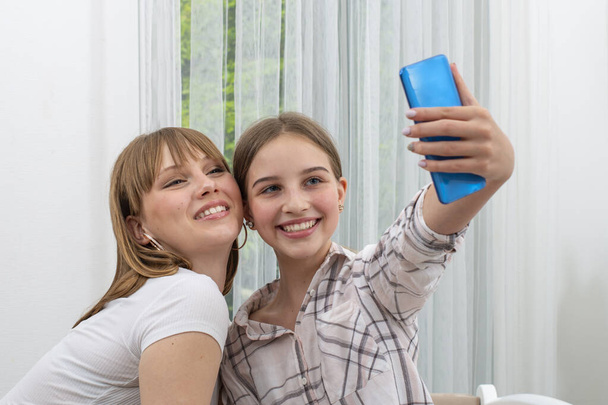 Two young girls  having fun resting at home spends free time together,  female holds smartphone make selfie photo with girlfriends smiling posing recording video for social networkin - Photo, image