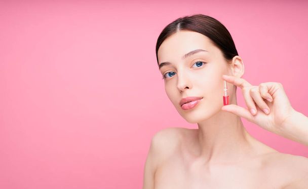 A nude woman holding an ampoule of serum for hair or skin care. Photo of an attractive woman with perfect natural makeup on a pink background. Beauty concept. Copy space. - Foto, imagen