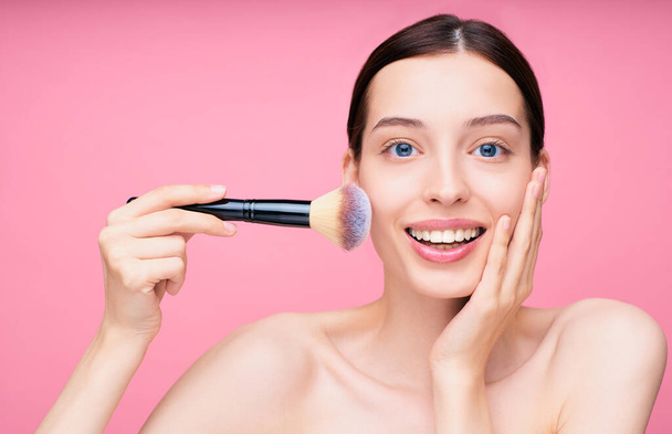 An image of a young beautiful Caucasian semi-nude woman looking at the camera and holding a makeup brush, isolated on a pink background. Make-up concept, makeup brushes, beauty and naturalness. - Foto, imagen
