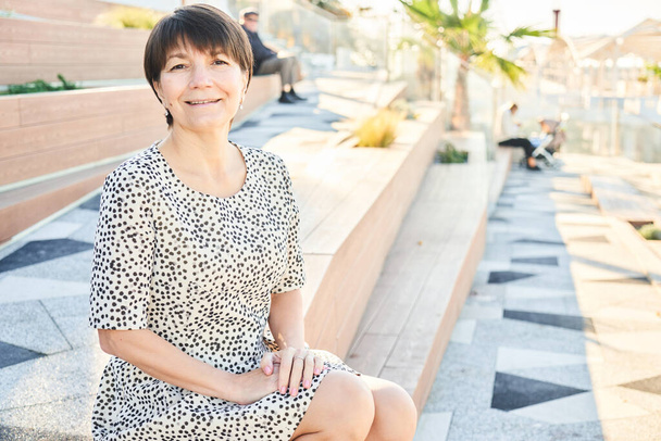 Portrait of 50 year old happy caucasian woman sitting and smiling in the park on the bench, adult healthy real lady - 写真・画像