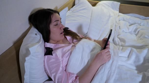 Frau liegt im Bett hält Smartphone liest E-Book schläft ein - Filmmaterial, Video