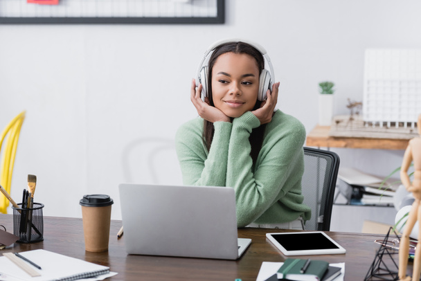 diseñador de interiores afroamericano feliz en los auriculares sentados en el lugar de trabajo, borrosa primer plano - Foto, Imagen