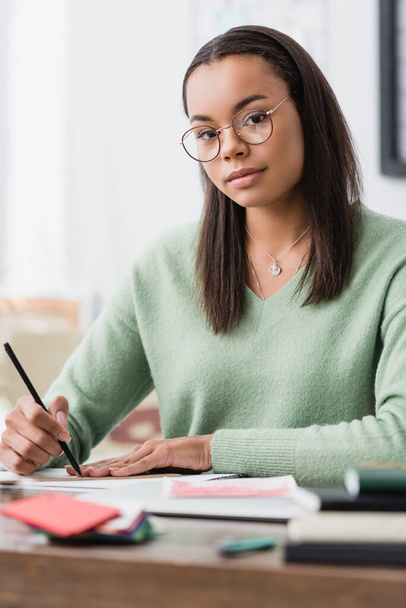 african american interior designer in eyeglasses looking at camera while holding pencil at workplace, blurred foreground - Fotó, kép