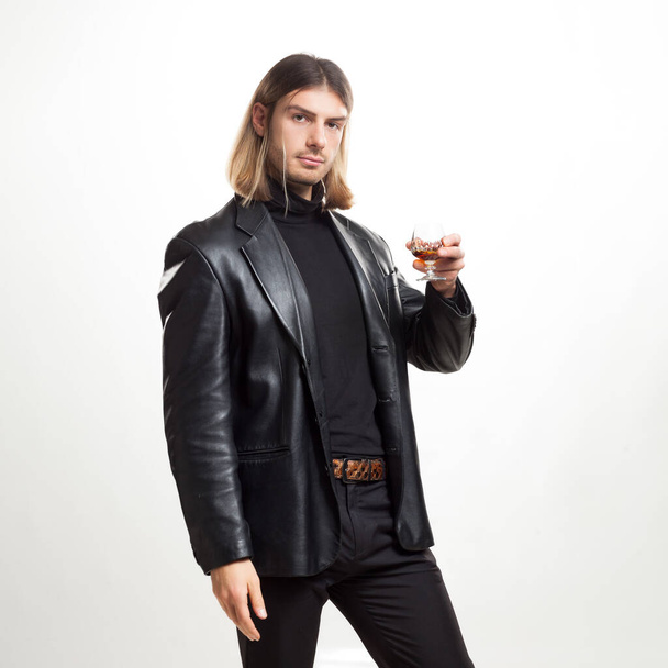 Портрет молодого человека светлые волосы со стаканом бренди в черном костюме, позирующего на сером фоне. Классический стиль. Снимок студии - Фото, изображение